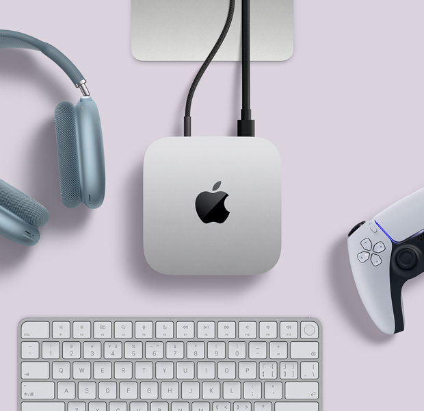 银色 Mac mini 的俯视图,两根连接线插入背面端口,设备旁边摆放着蓝色 AirPods Max、银色妙控键盘和无线游戏手柄。